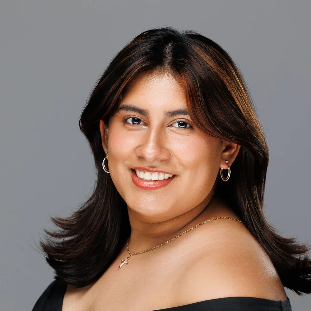 Headshot of a smiling woman with dark wavy hair in a black off-shoulder top on a gray backdrop