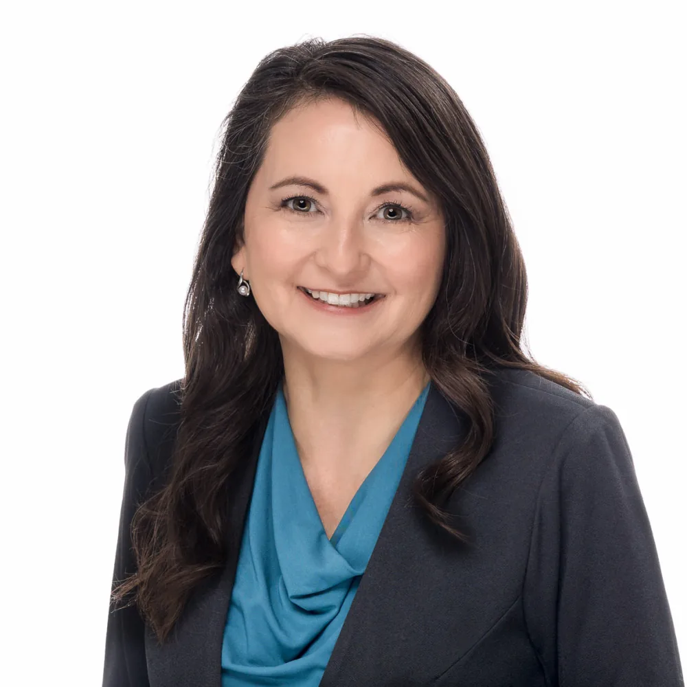 Professional headshot of a smiling woman with long dark hair in a charcoal blazer and teal blouse on a white backdrop